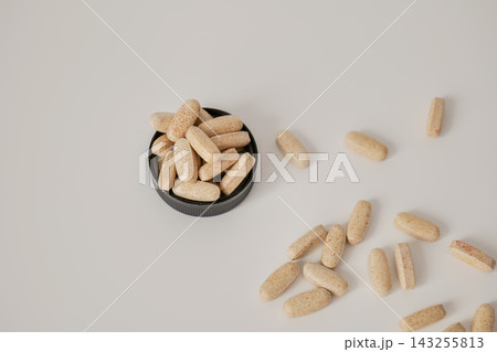 Beige capsules filling a black round lid with loose capsules scattered on a white surface. Capsule quantity and supplement organization concept Beige capsules filling a black round lid with loose capsules scattered on a white surface. Capsule quantity and supplement organization concept 143255813