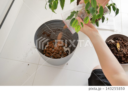 Two hands pour fresh brown soil or plant material from a bag into a gray plastic planter on a white tiled floor. Potting and repotting process Two hands pour fresh brown soil or plant material from a bag into a gray plastic planter on a white tiled floor. Potting and repotting process 143255850