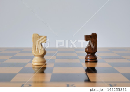 Light and dark chess knights face each other on a glossy wooden board. The minimal duel represents competition, confrontation, and strategic opposition 143255851