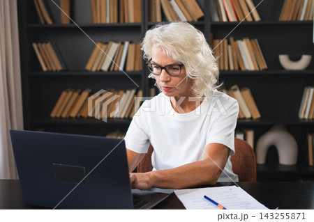 Confident stylish middle aged senior woman using laptop at workplace. Older mature lady businesswoman sitting at office table with laptop and paper document. Boss leader teacher professional worker 143255871