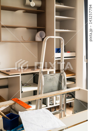 Setting up wooden cabinetry at a home improvement site using a step ladder for reach. Workspace features multiple shelves, tools, and building materials visible. New kitchen concept.  143257794