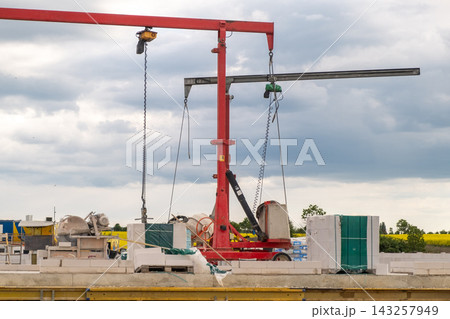 Heavy-duty crane positioned at a construction site, lifting concrete blocks and other materials. Various construction elements such as pallets and mixers are visible in the background, highlighting an 143257949