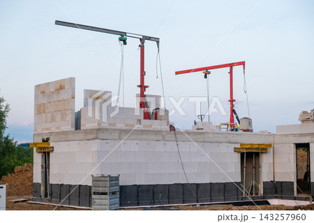 Construction cranes positioned above a residential building site, lifting heavy block materials. The structure features concrete walls and an organized workspace with construction materials visible. Construction cranes positioned above a residential building site, lifting heavy block materials. The structure features concrete walls and an organized workspace with construction materials visible. 143257960