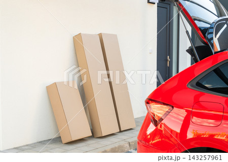 Large cardboard delivery boxes positioned against a wall next to a parked red vehicle. Clear sky and modern residential setting visible in the background. 143257961
