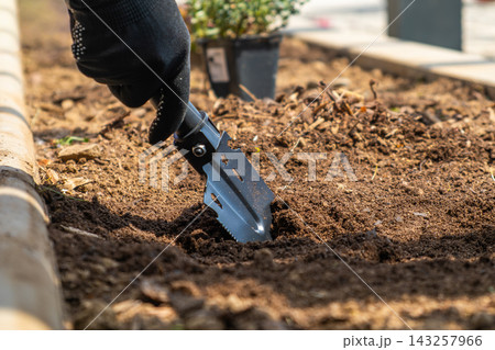Garden trowel being used to cultivate loose soil in a flower bed during gardening. A black glove-clad hand operates the tool, with a potted plant visible in the backdrop.  143257966