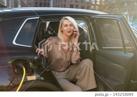 Engaging with a mobile phone, a blonde woman sits at the open door of an electric SUV during charging. The background features modern buildings and natural daylight streaming through. Engaging with a mobile phone, a blonde woman sits at the open door of an electric SUV during charging. The background features modern buildings and natural daylight streaming through. 143258095