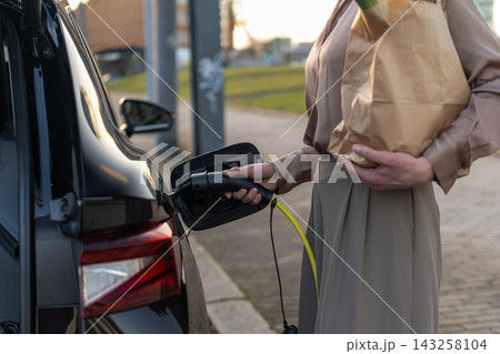 Female engaging electric vehicle charge port with charging cable while holding grocery bag. Outdoor urban setting with modern architecture and evening light creating a relaxed atmosphere. Female engaging electric vehicle charge port with charging cable while holding grocery bag. Outdoor urban setting with modern architecture and evening light creating a relaxed atmosphere. 143258104