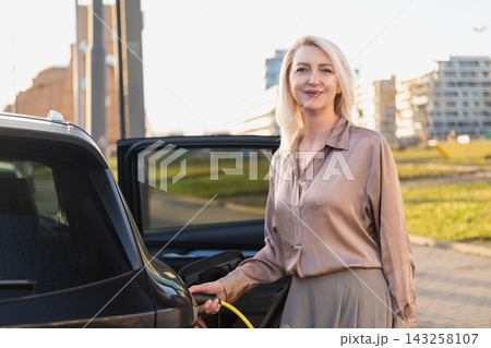 Electric vehicle blonde woman plugging in charging cable into car at urban parking area. Bright daylight with modern buildings visible in the background, providing a contemporary city setting. 143258107