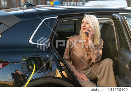 Businesswoman professional engaged in a phone conversation while seated in an electric vehicle at a charging station. The luxurious black SUV is plugged into the charger, showcasing modern technology. Businesswoman professional engaged in a phone conversation while seated in an electric vehicle at a charging station. The luxurious black SUV is plugged into the charger, showcasing modern technology. 143258109