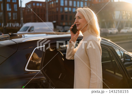 A professional woman engages in a phone conversation while exiting a sleek black sedan. The urban environment features modern buildings and soft sunlight illuminating the scene. A professional woman engages in a phone conversation while exiting a sleek black sedan. The urban environment features modern buildings and soft sunlight illuminating the scene. 143258155