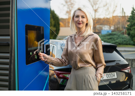Blonde woman engaging with electric vehicle charging station control panel in outdoor setting. Modern car parked nearby with clear sky and greenery visible in background. Blonde woman engaging with electric vehicle charging station control panel in outdoor setting. Modern car parked nearby with clear sky and greenery visible in background. 143258171