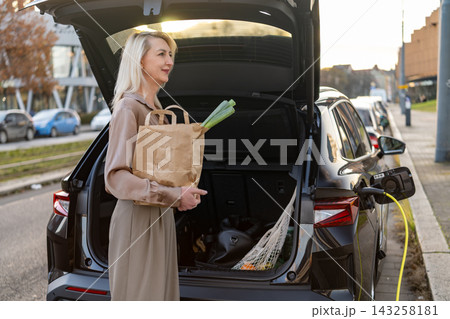 An electric car is being charged, a shopper removes a paper bag filled with fresh produce from the trunk. An electric car is being charged, a shopper removes a paper bag filled with fresh produce from the trunk. 143258181