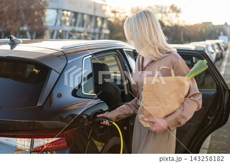 Charging an electric vehicle with a charging cable in a busy parking area. A woman holds a paper bag containing groceries and a green vegetable as she prepares to plug in the car. Charging an electric vehicle with a charging cable in a busy parking area. A woman holds a paper bag containing groceries and a green vegetable as she prepares to plug in the car. 143258182