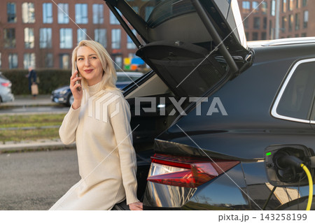 Engaging with a smartphone, a woman sits on the rear edge of an electric car during charging. Urban environment features modern buildings and vehicles visible in the background. Engaging with a smartphone, a woman sits on the rear edge of an electric car during charging. Urban environment features modern buildings and vehicles visible in the background. 143258199