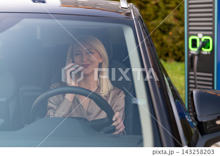 Blonde female driver using mobile phone while parked at electric vehicle charging station. Cleanly designed charging unit and green indicator light visible in the background, showcasing an eco 143258203