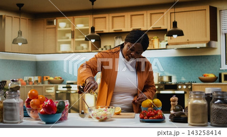 Chef vlogger using lemon juice and olive oil for seasoning the salad during filming, easy homemade recipe under natural light. Black woman showcasing meal prep and culinary development. Chef vlogger using lemon juice and olive oil for seasoning the salad during filming, easy homemade recipe under natural light. Black woman showcasing meal prep and culinary development. 143258414