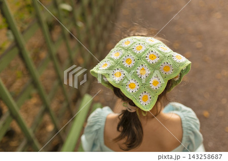 Back view of young girl wearing beautiful handmade green crochet headscarf with white daisy flowers. Cozy summer fashion accessory for kids, cottage core aesthetic, and handcrafted apparel concept  143258687