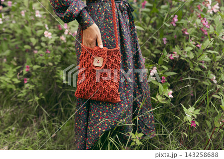 Close up of woman wearing a dark vintage floral dress and holding handmade terracotta orange crochet shoulder bag with wooden button. Standing in green meadow with wild pink flowers. Cottagecore Close up of woman wearing a dark vintage floral dress and holding handmade terracotta orange crochet shoulder bag with wooden button. Standing in green meadow with wild pink flowers. Cottagecore 143258688