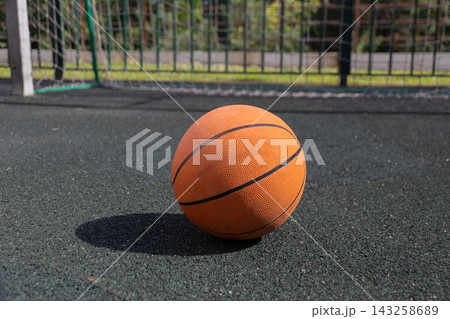 Close up of classic orange basketball on black rubber outdoor sports court surface. In the blurred background is soccer or handball goal net on sunny summer day. Street sports concept 143258689