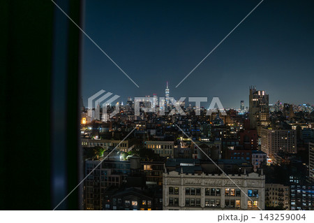 Wide panoramic view of New York City skyline at night with One World Trade Center glowing in Lower Manhattan. The urban cityscape is framed by nearby buildings and rooftops, highlighting illuminated Wide panoramic view of New York City skyline at night with One World Trade Center glowing in Lower Manhattan. The urban cityscape is framed by nearby buildings and rooftops, highlighting illuminated 143259004