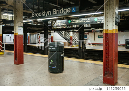 Wide view of Brooklyn Bridge City Hall subway station platform in New York City. Black litter bin stands on the platform beneath directional transit signage and fluorescent lighting. Wide view of Brooklyn Bridge City Hall subway station platform in New York City. Black litter bin stands on the platform beneath directional transit signage and fluorescent lighting. 143259033