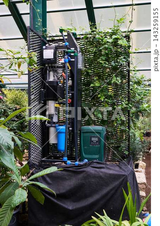 Glassed greenhouse irrigation system featuring pump, bundled hoses, blue filter, valves, metal frame, vertical piping, and climbing vines creating functional cultivated backdrop inside glass enclosure 143259155