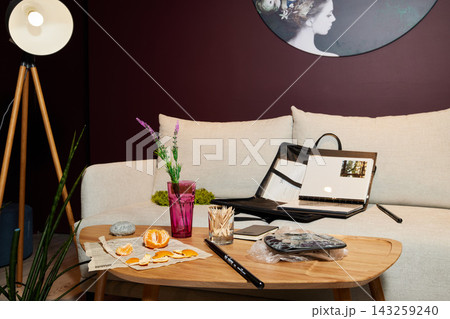 Living room scene with beige sofa, tripod wooden floor lamp, purple wall, round coffee table, open magazine, pink glass vase, orange peels on newspaper, desk accessories Living room scene with beige sofa, tripod wooden floor lamp, purple wall, round coffee table, open magazine, pink glass vase, orange peels on newspaper, desk accessories 143259240