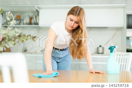 Portrait of focused teenage girl in casual wear wiping dust on table with rag while cleaning modern kitchen indoors Portrait of focused teenage girl in casual wear wiping dust on table with rag while cleaning modern kitchen indoors 143259728