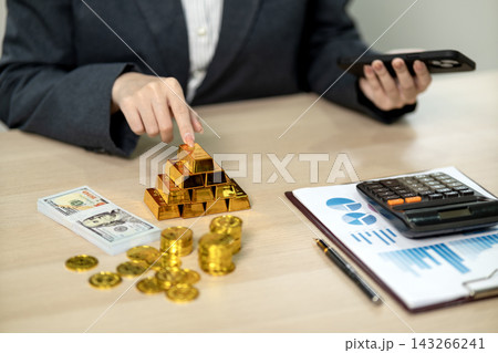 Businessperson Touching Gold Bar Pyramid While Holding Smartphone at Office Desk with Cash and Financial Documents Businessperson Touching Gold Bar Pyramid While Holding Smartphone at Office Desk with Cash and Financial Documents 143266241