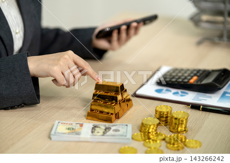 Businesswoman Touching Stacked Gold Bars While Using Smartphone at Office Desk with Cash and Coins 143266242