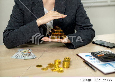 Businesswoman Protecting Stack of Gold Bars with Hands at Office Desk with Money and Calculator Businesswoman Protecting Stack of Gold Bars with Hands at Office Desk with Money and Calculator 143266555
