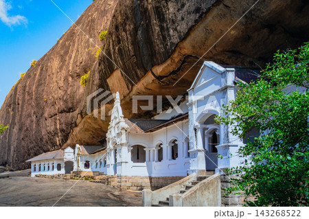 Dambulla Cave Temple in Sri Lanka beneath giant rock formation with ancient Buddhist monastery architecture and historic courtyard. Famous UNESCO heritage and cultural travel destination Dambulla Cave Temple in Sri Lanka beneath giant rock formation with ancient Buddhist monastery architecture and historic courtyard. Famous UNESCO heritage and cultural travel destination 143268522