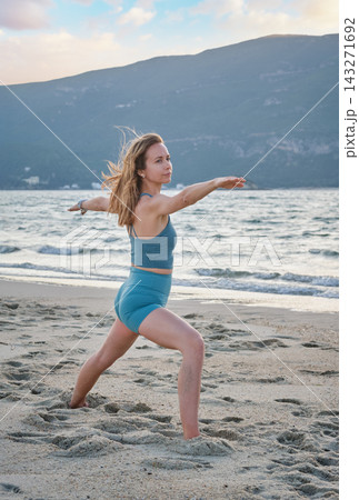 A woman stands on beach sand in Virabhadrasana 2 yoga, Warrior 2 Pose, with a bent front knee and arms extended sideways as she gazes along her forward hand 143271692