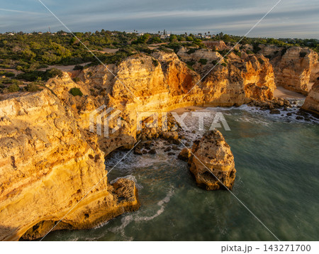 Drone view of Praia da Marinha in Algarve, Portugal, with golden limestone cliffs, a small sandy cove and an offshore sea stack in soft sunrise light. Drone view of Praia da Marinha in Algarve, Portugal, with golden limestone cliffs, a small sandy cove and an offshore sea stack in soft sunrise light. 143271700