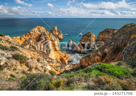 View from the clifftop path at Ponta da Piedade in Lagos, Algarve, Portugal, with golden limestone sea stacks, turquoise water and a wooden staircase leading down to the cove. 143271708