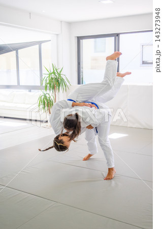 Two female athletes in white gis and blue belts practice grappling martial arts inside a bright studio, demonstrating a jiu jitsu hip throw takedown on the tatami mat Two female athletes in white gis and blue belts practice grappling martial arts inside a bright studio, demonstrating a jiu jitsu hip throw takedown on the tatami mat 143273948