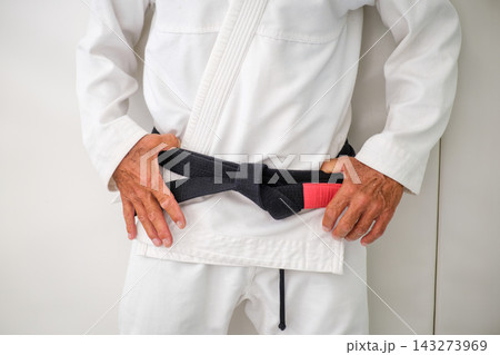 Close up of a tied Brazilian jiu jitsu black belt with a red rank bar over a white gi, with the hands of the practitioner beside the knot 143273969