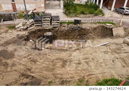 Urban excavation site reveals exposed earth, soil mounds, utility pipes, and concrete blocks with wooden pallets along cleared street edge beside apartment buildings 143274624