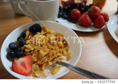 Bowl of cornflakes topped with fresh strawberries, blueberries, and blackberries, with a cup of coffee and assorted fruits on a wooden table 143277744