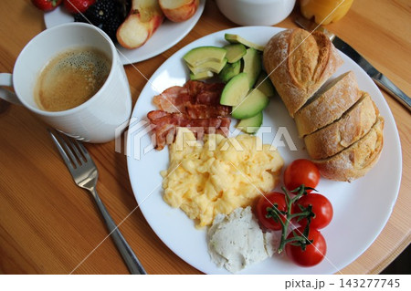 Breakfast plate featuring scrambled eggs, crispy bacon, sliced avocado, cherry tomatoes, fresh bread, and a cup of coffee on a wooden table 143277745