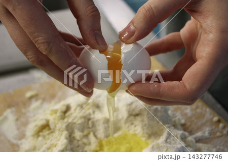 Hands breaking a fresh egg over a mound of flour on a wooden surface, with egg yolk and white spilling into the flour, kitchen setting visible in the background 143277746