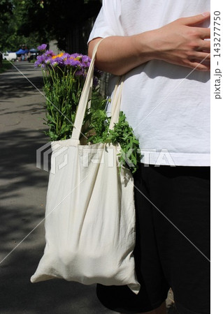 Individual holding a reusable cotton tote bag filled with fresh flowers and herbs while walking along a sunlit urban street with greenery in the background Individual holding a reusable cotton tote bag filled with fresh flowers and herbs while walking along a sunlit urban street with greenery in the background 143277750