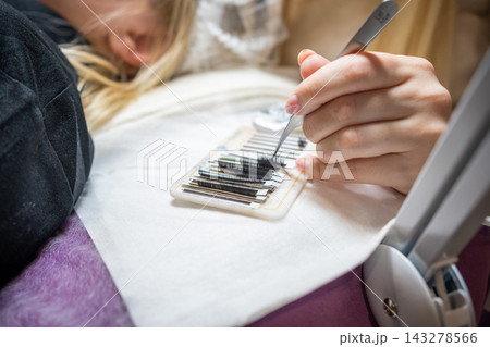 The master takes a bunch of curved artificial eyelashes with tweezers from the tablet and dips into the glue. The process of eyelash extension in a beauty salon.  143278566