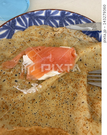 Savory crepe with cream cheese and smoked salmon on blue patterned plate, close up breakfast dish with fork and knife, rustic homemade brunch meal with soft colors and textured surface 143279360