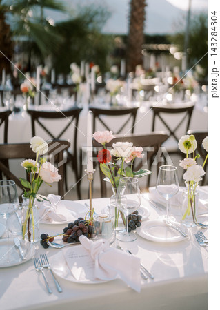 Elegant outdoor banquet table with white linen, floral bud vases, grapes, glassware, candle centerpiece, polished cutlery, dark chairs, and soft sunlit tropical garden backdrop Elegant outdoor banquet table with white linen, floral bud vases, grapes, glassware, candle centerpiece, polished cutlery, dark chairs, and soft sunlit tropical garden backdrop 143284304
