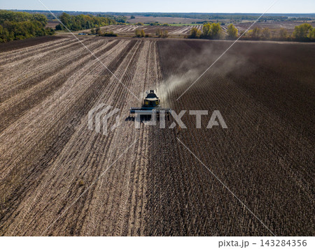 Combine harvester crosses sunflower field, harvesting mature rows and leaving parallel strips of residue. Farm machine raises visible dust cloud across expansive farmland under clear sky. Combine harvester crosses sunflower field, harvesting mature rows and leaving parallel strips of residue. Farm machine raises visible dust cloud across expansive farmland under clear sky. 143284356