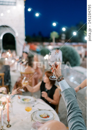 Evening celebration outdoors. Guests toast wine under lighting. Outdoor dinner with friends and festive decor Evening celebration outdoors. Guests toast wine under lighting. Outdoor dinner with friends and festive decor 143284795