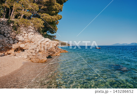 Scenic coastal view featuring clear turquoise water, rocky shoreline, and lush green trees under a bright blue sky in a serene natural environment 143286652