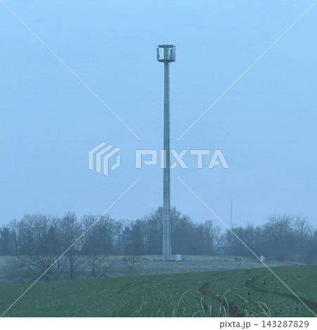 Tall cellular communication tower standing in a misty rural landscape with green agricultural fields and leafless trees on a winter morning 143287829