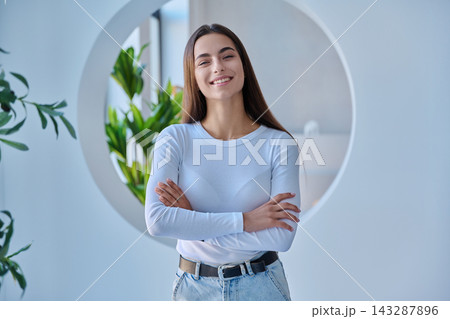 Smiling young female student with crossed arms standing in modern office or campus setting Smiling young female student with crossed arms standing in modern office or campus setting 143287896
